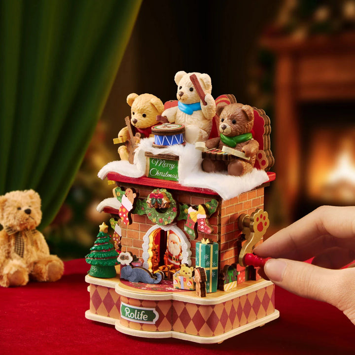 Hand turning the red crank handle on the side of the Rolife Christmas fireplace music box, with teddy bears on top and a plush bear beside it on red fabric.