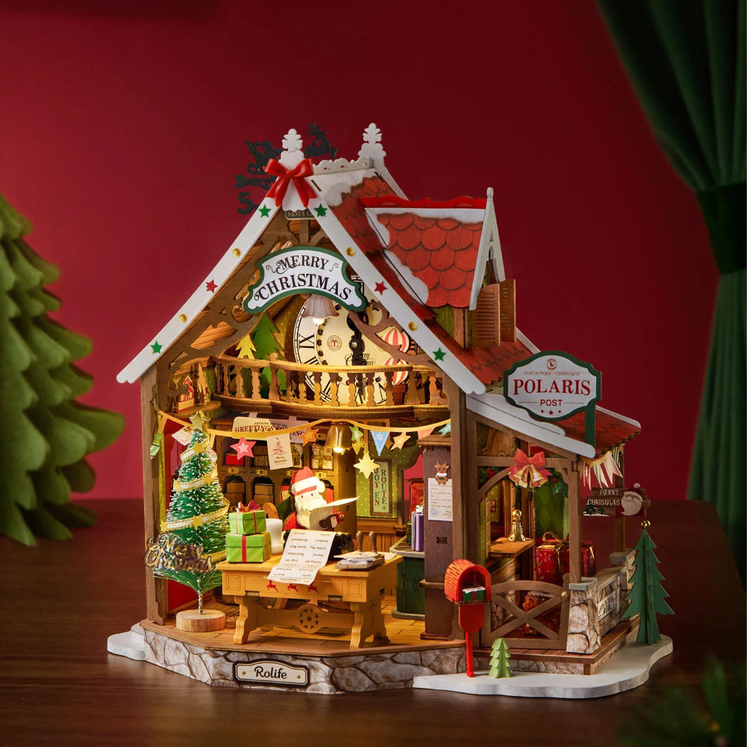 Rolife wooden miniature Christmas post office with red roof, Santa figure at desk, decorated tree, red mailbox and Polaris Post sign, lit warmly against a red backdrop.