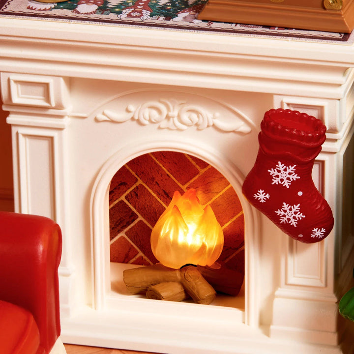 Close-up of a miniature white fireplace with a glowing orange flame, logs and a red snowflake stocking.