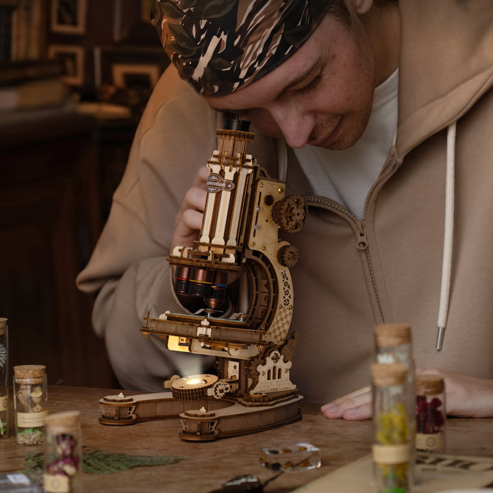 Person in a beige coat leaning over the ROKR wooden microscope on a vintage desk with small glass vials and warm ambient light
