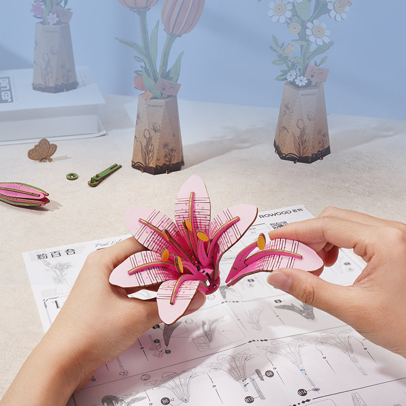 Hands assembling a Rowood Pink Lily 3D wooden flower puzzle, holding the bright pink and magenta lily bloom with yellow stamens above the instruction sheet and other flower components